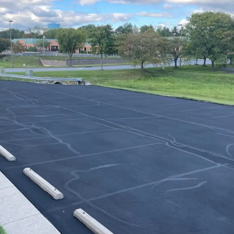 An empty, freshly paved black asphalt parking lot with concrete barriers, surrounded by greenery and buildings in the background.