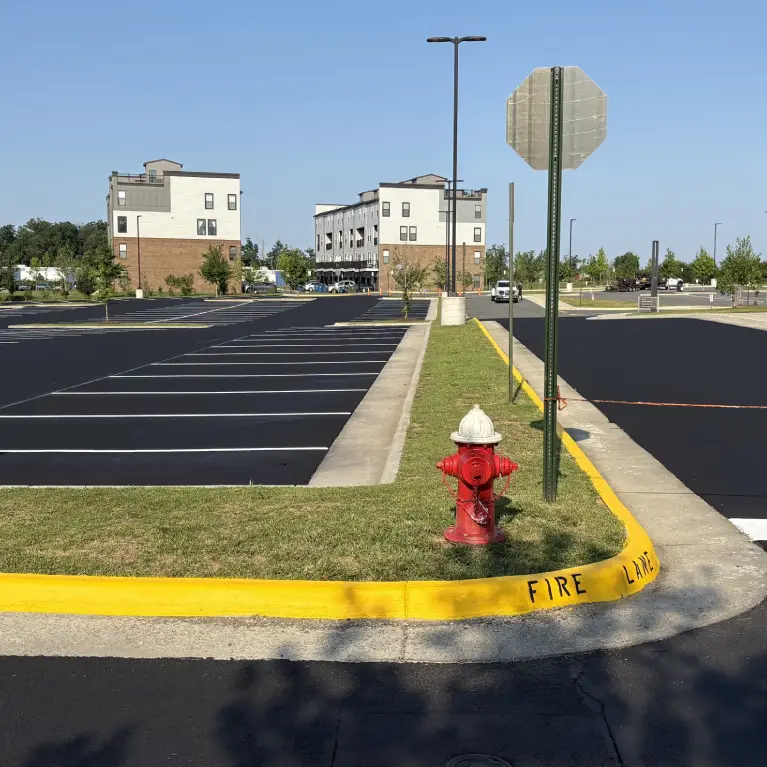 A newly paved parking lot with empty spaces, a red fire hydrant, and two multi-story buildings in the background under a clear blue sky.